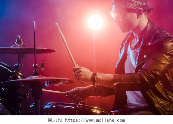 在蓝红色灯光下的帅气鼓手年轻的男性音乐家在舞台上的摇滚音乐会打鼓皮夹克的侧视图  